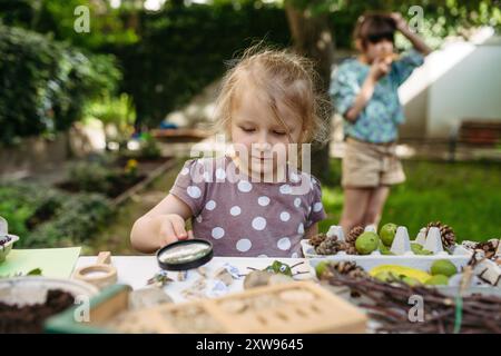 Little girl looking at models of insect with magnifying glass, learning about wildlife in forest kindergarten. Stock Photo