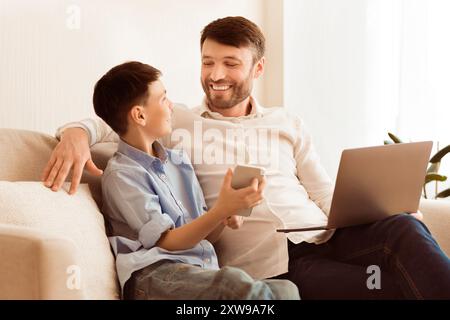 Father And Son Using Phone And Laptop Sitting At Home Stock Photo