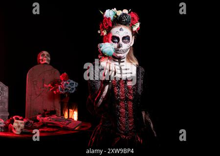 Portrait of woman holding cyan colored rose during Day of the Dead Halloween event honoring dead loved ones. Scary lady acting as devil witch during ancient Aztec custom of celebrating ancestors Stock Photo
