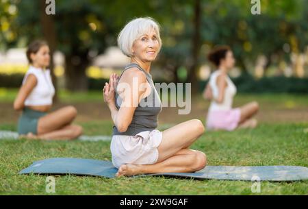 Elderly woman performing Reverse Prayer Gomukhasana during group yoga in park Stock Photo