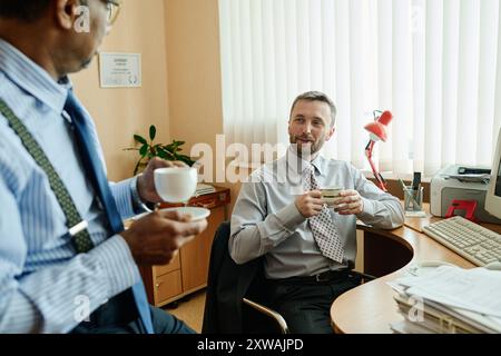 Two colleagues having coffee while discussing ideas at workstation. Both are engaged in casual conversation with coffee cups in hand adding relaxed ambiance to office Stock Photo