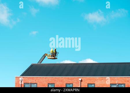 Roofers working off telescopic boom lift while inspecting and replacing roof tiules on a  multistorey residential building against blue sky with space Stock Photo