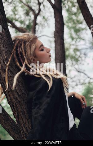 Woman with blonde dreadlocks leans against a tree in a forest, wearing a black jacket and white shirt, while looking off to the side. Stock Photo