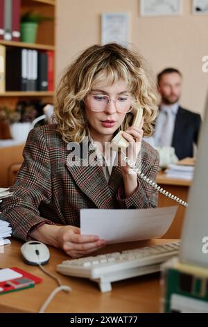 Woman with curly blond hair wearing glasses and plaid blazer talking on phone while holding document sitting at desk in office, man with beard working in background Stock Photo