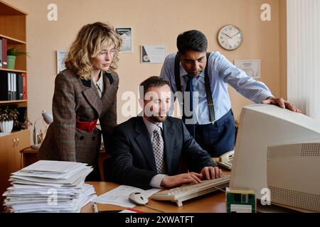 Three professionals in office discussing documents displayed on computer monitor, with one person seated and two standing around desk Stock Photo