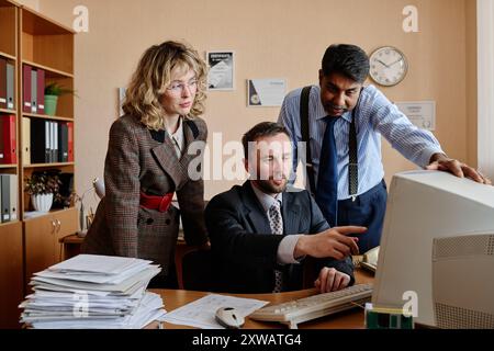 Three colleagues of diverse backgrounds collaborating on business project in office setting with computer and paperwork present on desk Stock Photo