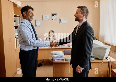 Two businessmen shaking hands in modern office setting with woman working in background, with documents and computer equipment present on desks creating professional environment Stock Photo