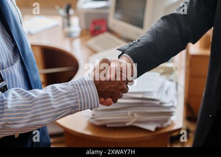 Two professionals completing business handshake in corporate office environment with paperwork on table, symbolizing agreement and partnership between individuals in business Stock Photo