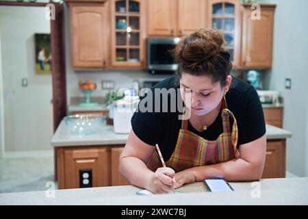 Woman writing notes in kitchen with phone and notepad. Stock Photo