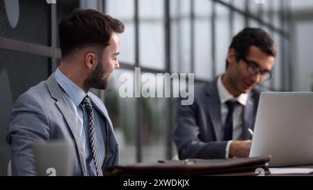 Two serious men in suits sitting at desk with laptop computer and talking. Stock Photo
