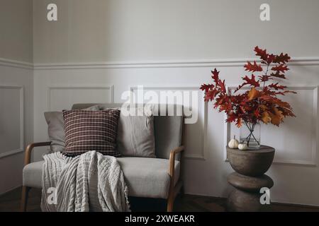 Red oak tree leaves, branches in vase on modern curved wooden side table. Linen midcentury sofa with throw blanket, checkered cushions. Little white Stock Photo