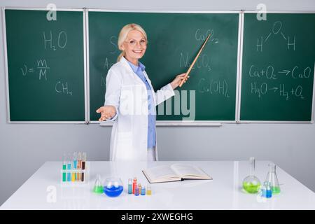 Photo of charming intelligent senior woman university lecturer in white coat explaining subject isolated on green board background Stock Photo