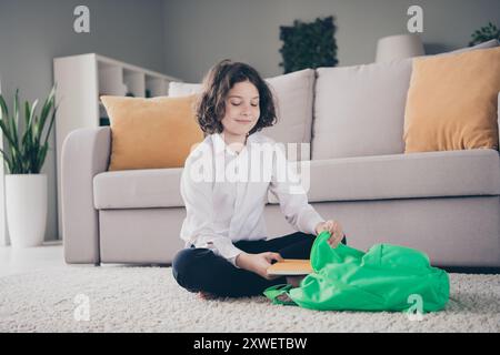 Photo of charming boy schoolchild preparing to school putting book in schoolbag indoors house living room Stock Photo