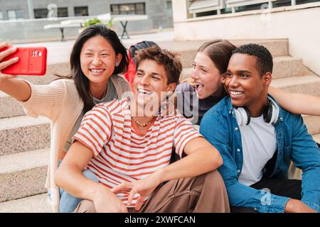Group of international students taking a selfie portrait sitting on the university campus stairway. Young real students having fun shooting a photo portrait with a cellphone for the social media. High quality photo Stock Photo