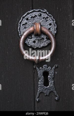 Heavy iron knocker, a little aged and rusted, set upon a patterned plate into a sturdy old wooden door. Keyhole below Stock Photo