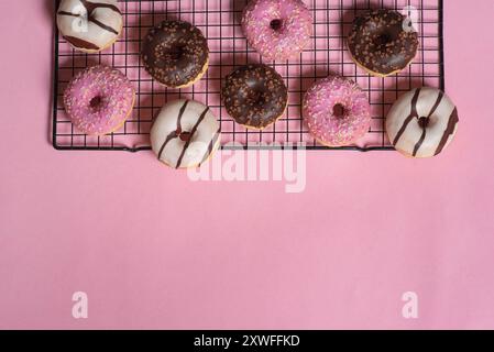 Image displays an assortment of donuts placed on a cooling rack with a pink background, showcasing a variety of toppings including sprinkles and choco Stock Photo