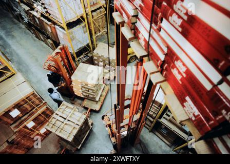 Two workers maneuver fork lifts around stacked pallets in a busy warehouse filled with boxes and storage racks. Stock Photo