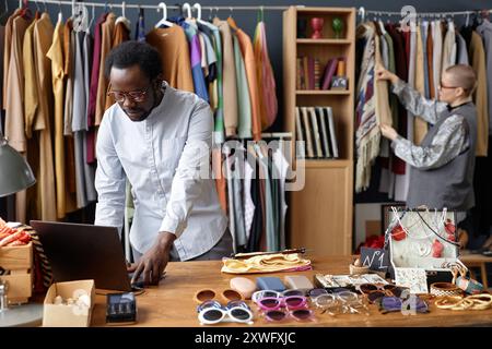 Medium shot of African American salesman using laptop on wooden table with fashion accessories for sale at thrift store checkout, copy space Stock Photo