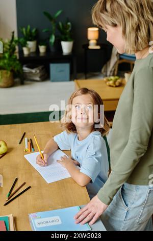 Blue eyed girl sitting at desk and looking at her mom while she standing next to her Stock Photo