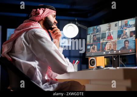 Detailed view of Arab entrepreneur attentively listening to his colleagues on a video call at his desk. Image showcasing group of people utilizing modern technology for online communication. Stock Photo