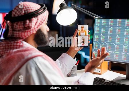 Middle Eastern entrepreneur having a serious discussion on financial reports with his colleagues on his smartphone in his home office. He wears Islamic clothing and has a professional demeanor. Stock Photo