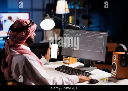 Male person in traditional attire sits at desk presenting a promo on mobile device with isolated copyspace template. The individual focus on phone reflects the integration of technology and culture. Stock Photo
