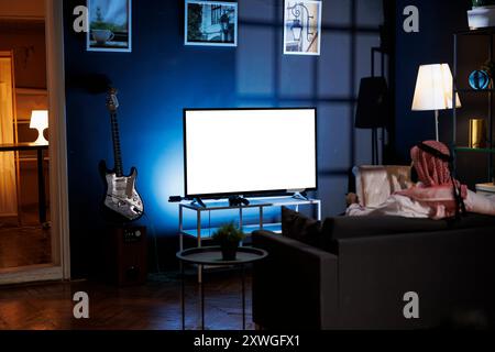Back-view shot of Muslim guy in traditional attire seated on sofa in front of television displaying isolated white screen. Image showing man looking at tv screen with blank copyspace mockup template. Stock Photo