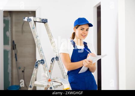 Positive young female builder contractor inspecting room and taking notes Stock Photo