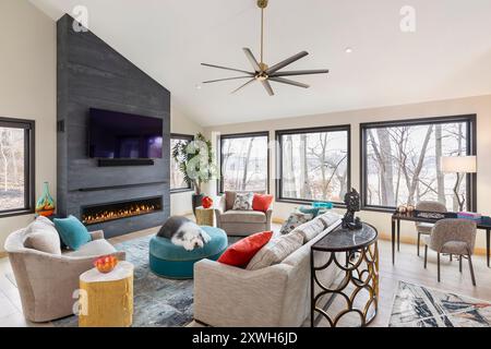 Angled view of living room with gray stone fireplace and large windows to outsdie with veiw of trees and large sheepdog on the ottoman Stock Photo