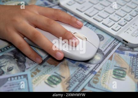 A close-up image of a hand holding a computer mouse, hovering over a bed of US dollar bills. Online business, pay per click, digital transaction Stock Photo