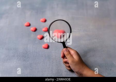 A hand holding a magnifying glass focuses on a single pink heart among a cluster of others. Quest for love concept. Stock Photo