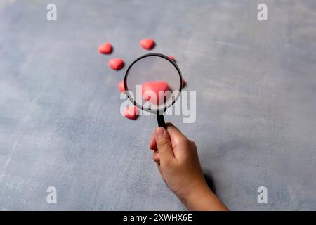 A hand holding a magnifying glass focuses on a single pink heart among a cluster of others. Quest for love concept. Stock Photo