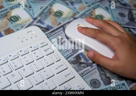 A close-up image of a hand holding a computer mouse, hovering over a bed of US dollar bills. Online business, pay per click, digital transaction Stock Photo