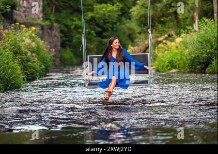 a smiling young woman swings on a rope swing across a fast-flowing river Stock Photo