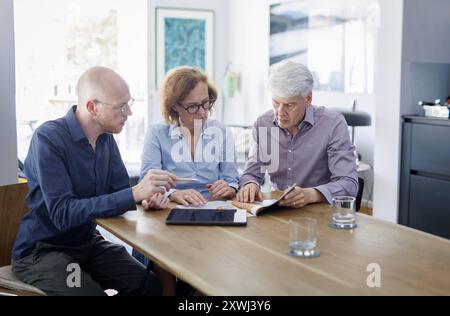 Symbolfoto zum Thema Beratung. Eine aeltere Frau und ein aelterer Mann sitzen zu Hause zusammen an einem Tisch und werden von einem jungen Mann berate Stock Photo