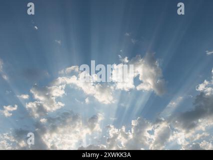 sun rays sweeping across a blue sky from behind clouds lighting up the high atmosphere haze Stock Photo