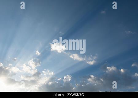 sun rays sweeping across a blue sky from behind clouds lighting up the high atmosphere haze Stock Photo