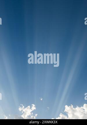 sun rays sweeping across a blue sky from behind clouds lighting up the high atmosphere haze Stock Photo