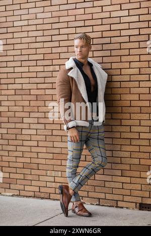 A handsome blonde man in a stylish brown jacket and plaid pants leans against a brick wall Stock Photo