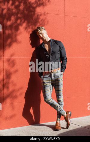 A handsome blonde man in stylish attire leans against a red wall Stock Photo