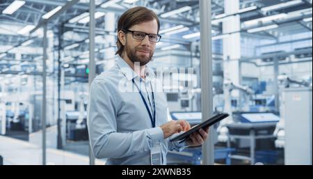 Portrait of a Handsome Adult Engineer Wearing Glasses, Using Tablet Computer and Looking at Camera. Technician in a Shirt Working in a Modern Technology Factory with Robotis Assembly Arms Stock Photo