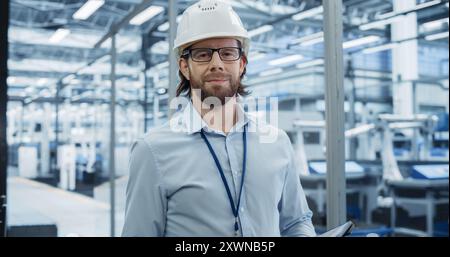 Young Electronics Manufacture Engineer Smiling, Looking at Camera. Portrait of a Man Wearing a Protective White Hard Hat. Specialist Monitoring Artificial Intelligence Systems at a Factory Stock Photo