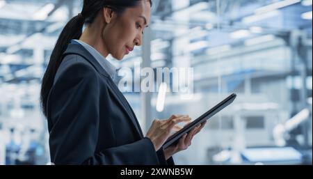 Beautiful Asian Female Engineer Standing Next to a Window in Office, Using Tablet Computer at an Electronics Manufacture. Specialist Using an Online Software to Program Robots on Assembly Line Stock Photo
