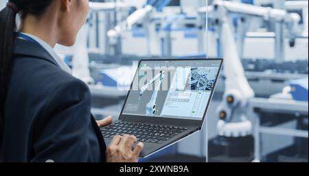 Manufacturing Robot Arms Administrator Programming Robotic Vision Systems to Execute Production at an Electronics Factory. Over the Shoulder Footage of a Female Supervising Assembly with a Computer Stock Photo
