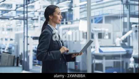 Production Line Technician Working on a Laptop Computer at Factory Office. Asian Female Software Specialist Updating Code for Machine Production and Assembly with Automated Robotic Arms Stock Photo