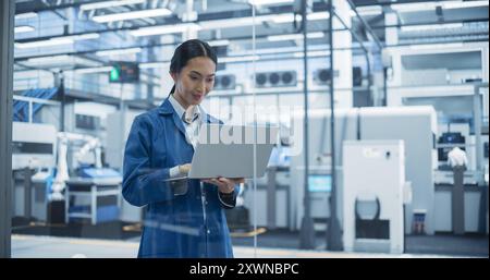 Portrait of a Beautiful Asian Engineer Using Laptop Computer and Looking Around a Factory Facility with Equipment Producing Modern Electronic Components with Robotic Arms on Assembly Conveyor Stock Photo