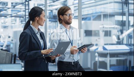 Factory Managers Using Computer Devices at Modern Manufacture with Autonomous Robot Hands. Female and Male Specialists Monitoring Technical Data, Analyzing Reports and Planning Updates Stock Photo