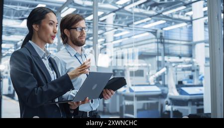 Two Multiethnic Engineers Standing in Office, Using Laptop and Tablet Computers at an Electronics Factory. Machines are Undergoing Maintenance, and Specialists Monitoring the Progress on Devices Stock Photo