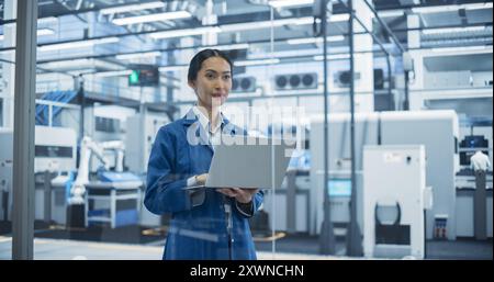 Electronics Manufacturing Technician in a Blue Coat Working on Laptop Computer at an Assembly Plant. Asian Female Specialist Developing, Maintaining, Setting Up Software for Automated Production Stock Photo