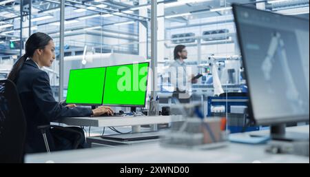Female Robotics Technician Working on Desktop Computer with Green Screen Chromakey Mock Up Display in a Factory Office. Technological Research and Development Facility with Autonomous Assembly Line Stock Photo